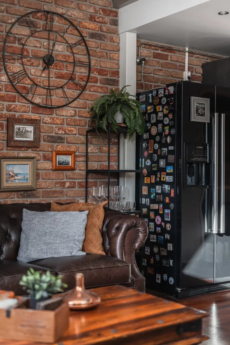 Iron wall clock on brick beside a fridge covered in travel magnets