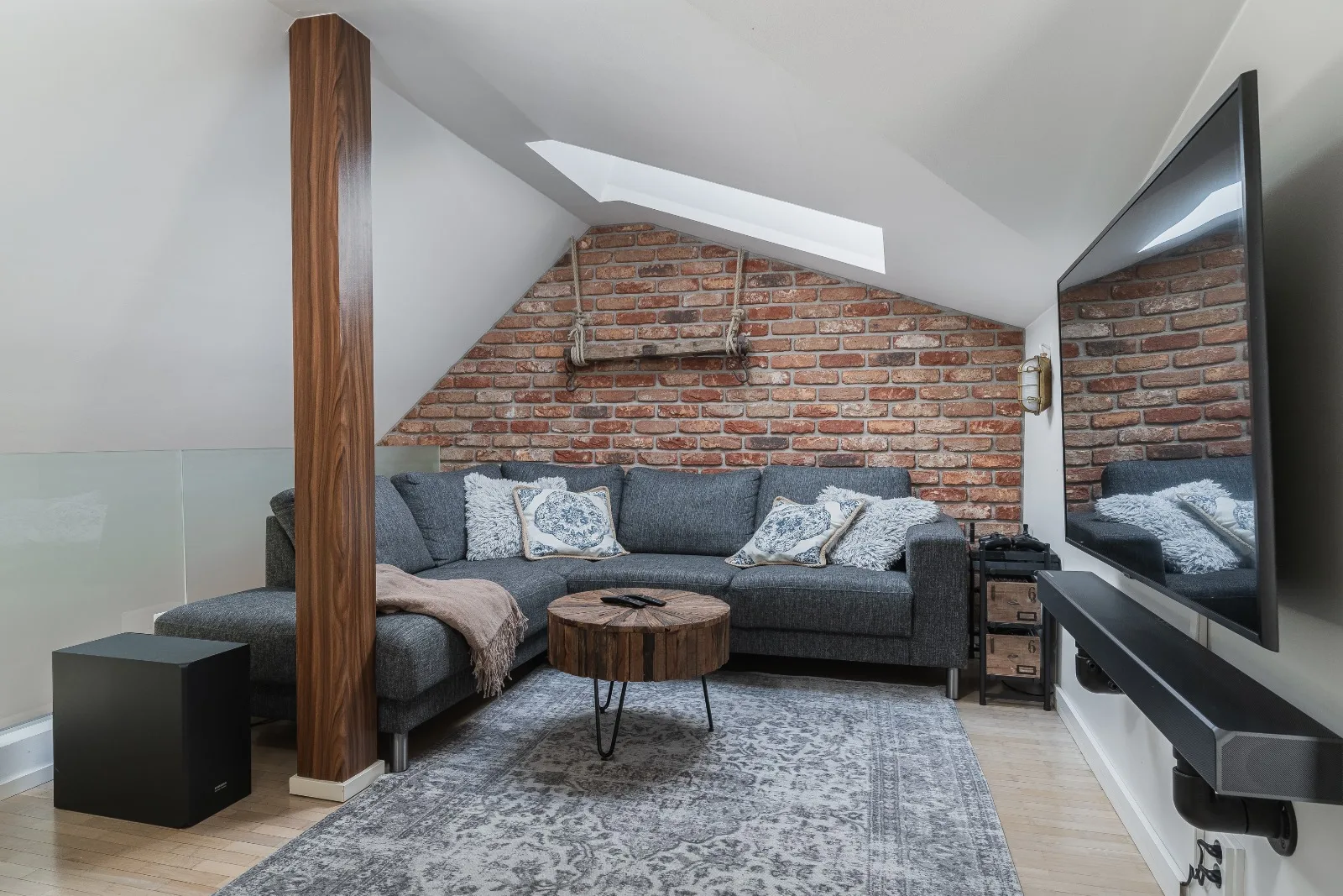 Upstairs mezzanine lounge with a grey corner sofa against a brick wall, a wall-mounted television and a skylight